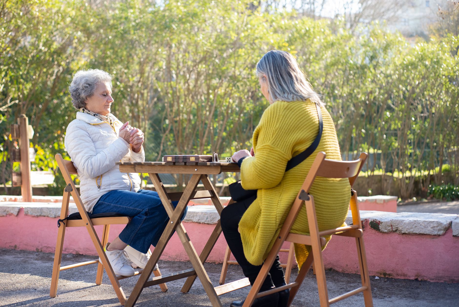 Senior women enjoying a game of dominoes at a wooden table in a sunny park setting.