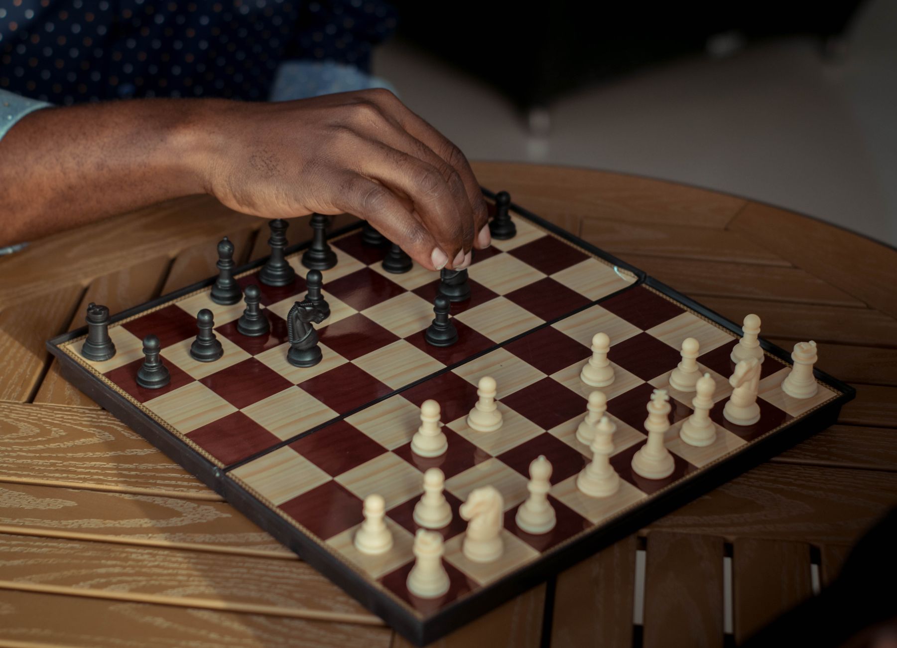 A close-up of a hand moving chess pieces on a board atop a wooden table.