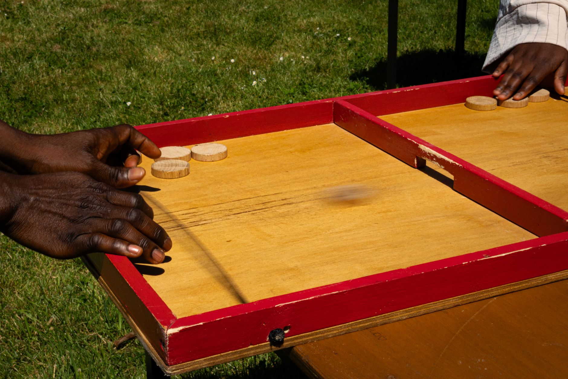 Hands playing a traditional wooden board game outdoors on a sunny day.