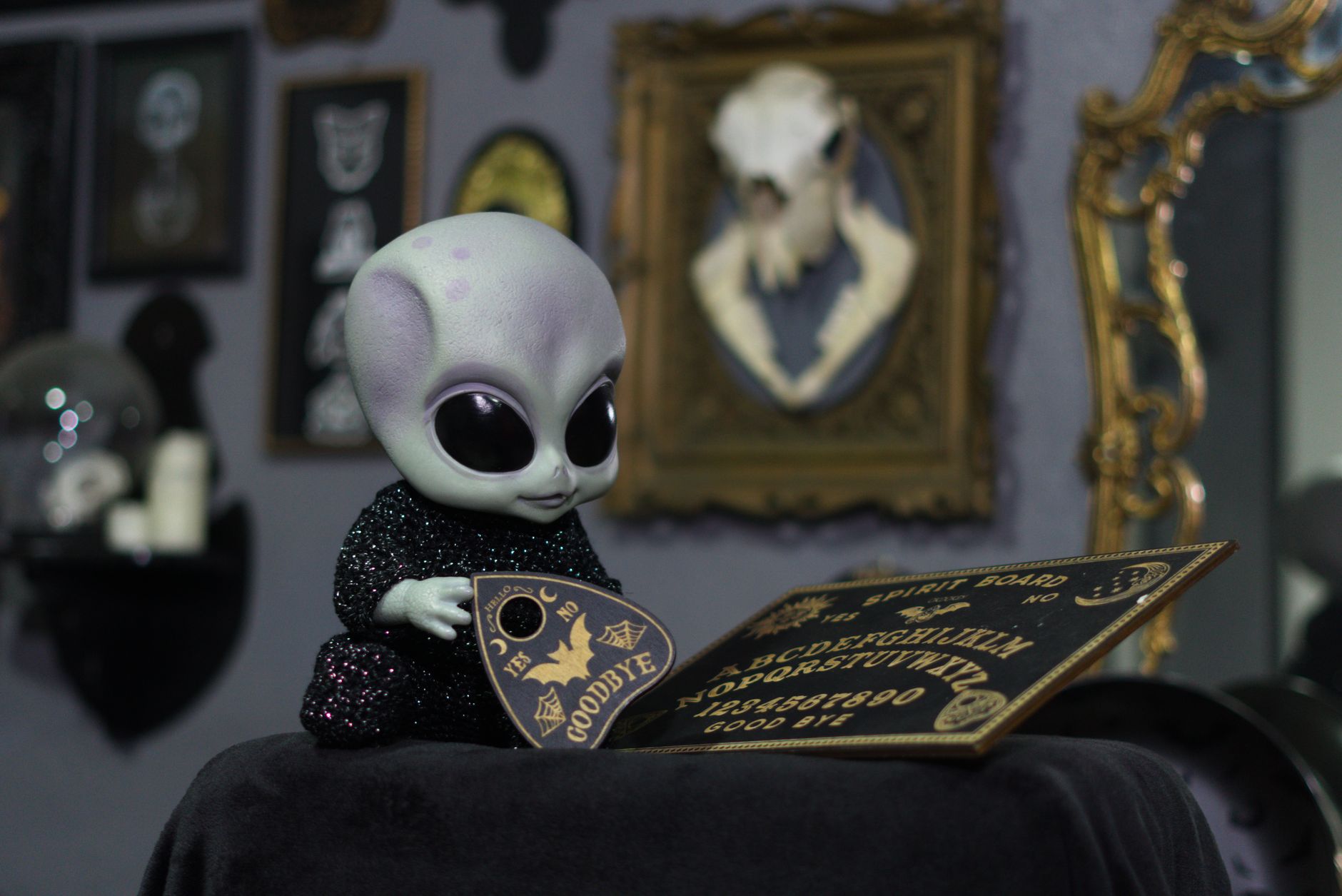 Alien doll interacting with an Ouija board in a decorated room with vintage frames.
