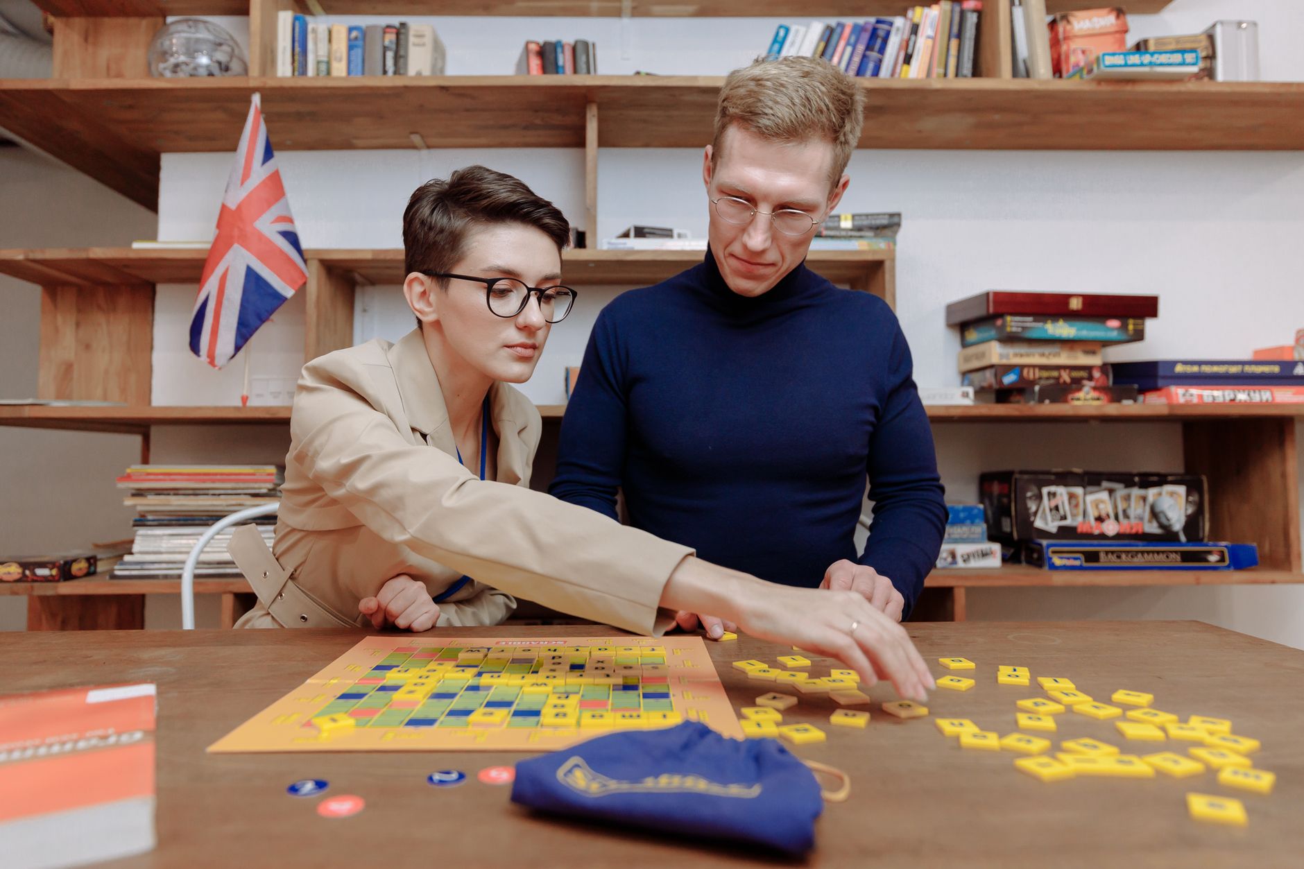 A man and woman play a board game together in a cozy library setting with books and a flag.