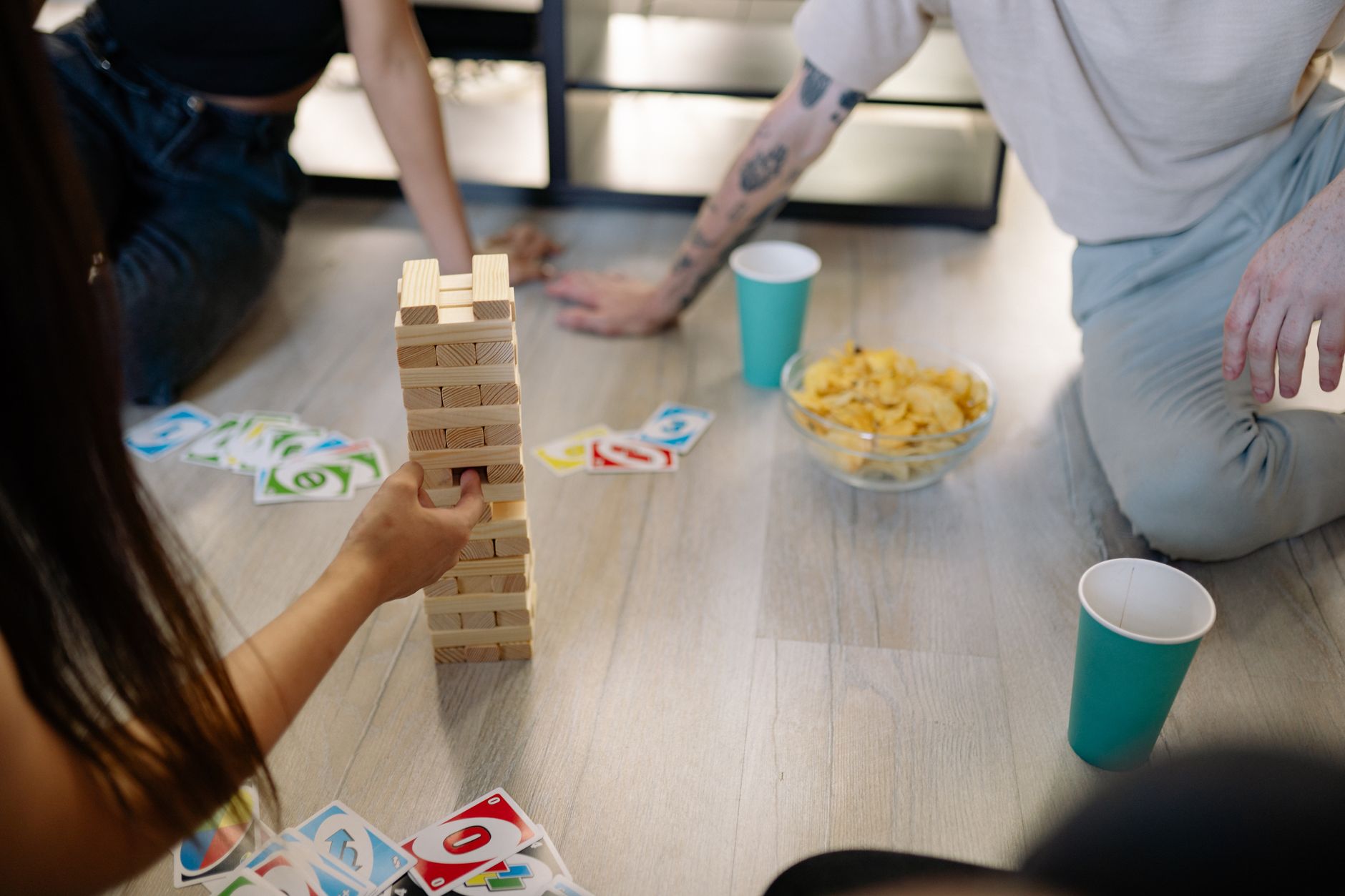 Group of friends playing Jenga and card games with snacks on wooden flooring indoors.