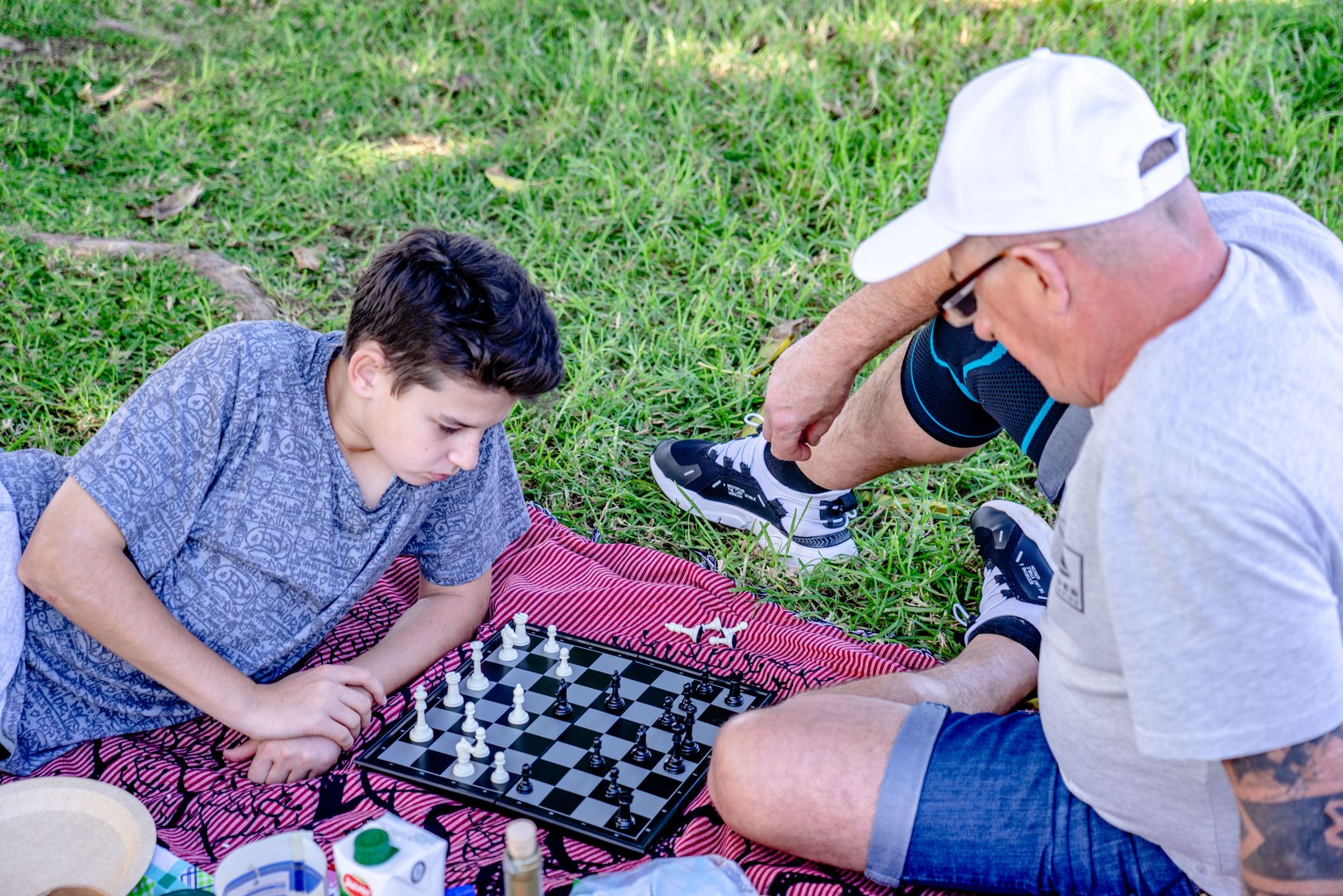A grandfather and grandson enjoy a chess game on a picnic blanket in a sunny park.