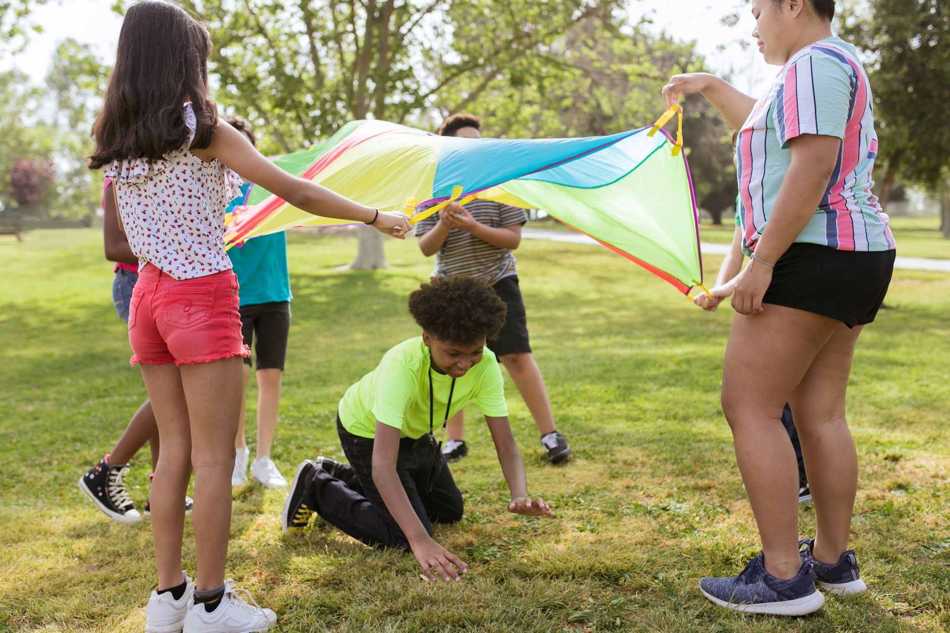 Group of children playing with a colorful parachute outdoors on a sunny day.