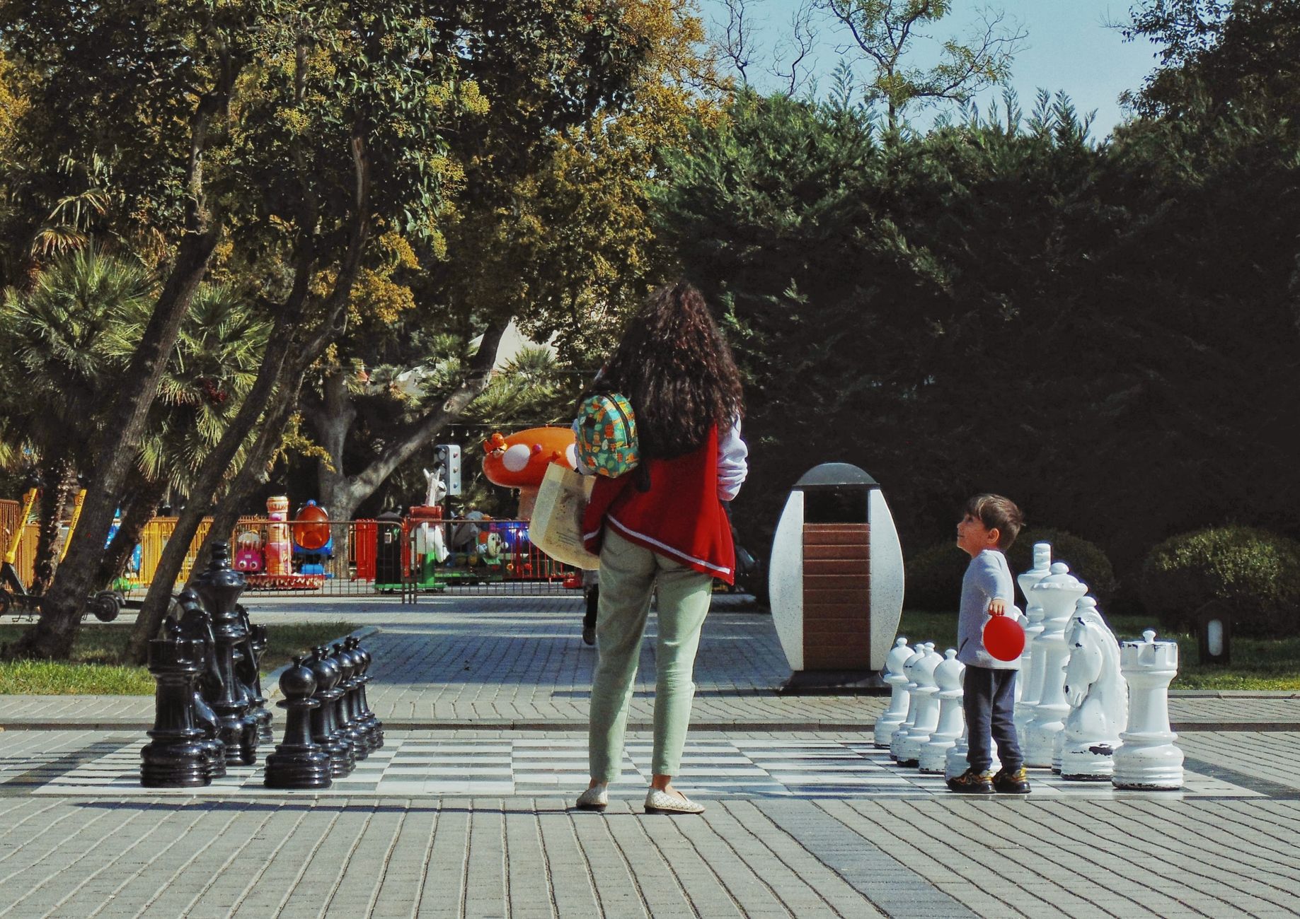 A woman and child engaged with a large outdoor chess game in a park setting.