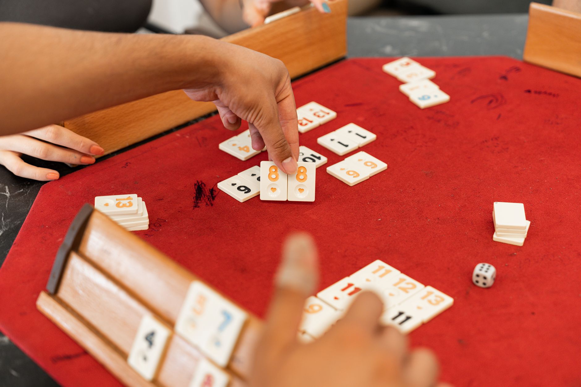 Close-up of hands playing Rummikub with numbered tiles on a red felt table.