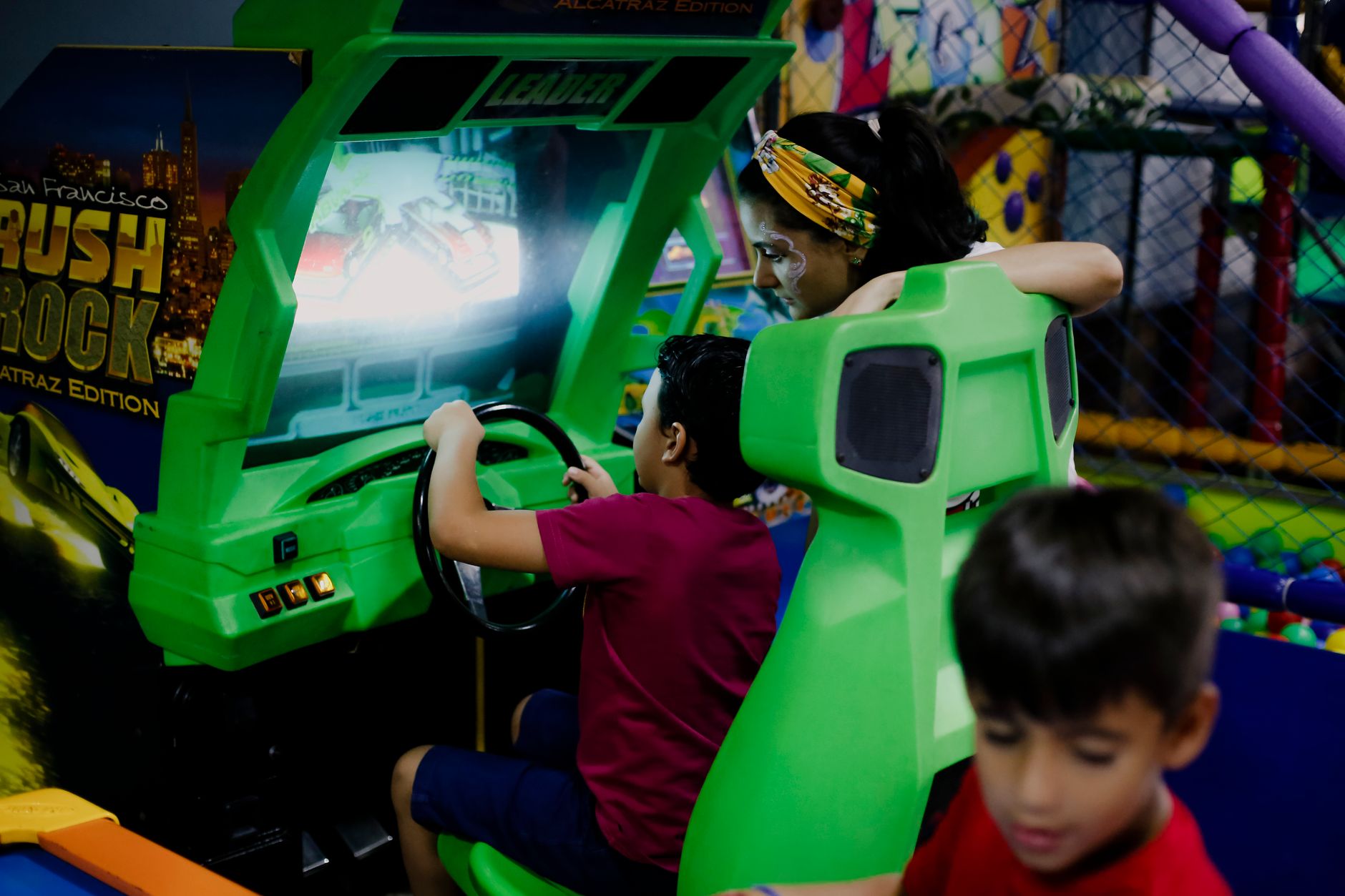Unrecognizable child having fun with bright car racing simulator while turning steering wheel near serious ethnic woman and funny brother in