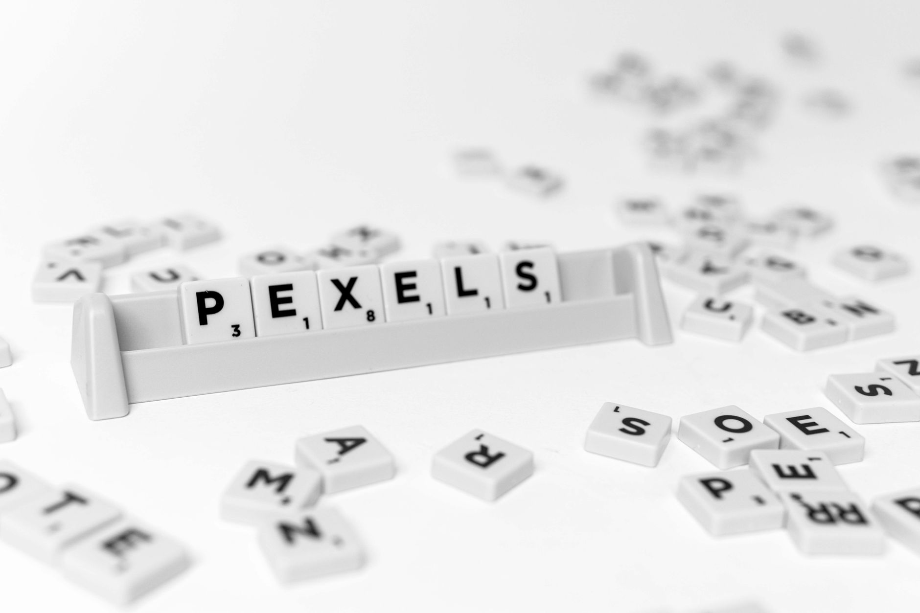 Monochrome image of Scrabble tiles spelling 'PEXELS' on a rack, scattered tiles in the foreground.